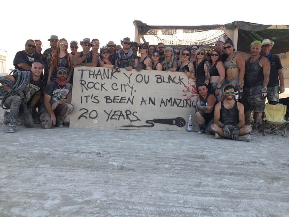 Photo of a group of dusty people standing behind and around a large plywood sign spray painted with a drawing of a dropped microphone and text reading: Thank you Black Rock City it's been an amazing 20 years. The foreground is caked playa and there is a large bottle of alcohol in front of the sign.