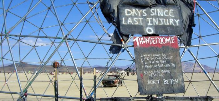 Photograph of the hand-painted Thunderdome Rules Sign on the side of a geodesic dome. In the background is flat playa with an art car, camps and mountains in the distance under a blue sky with small white clouds.